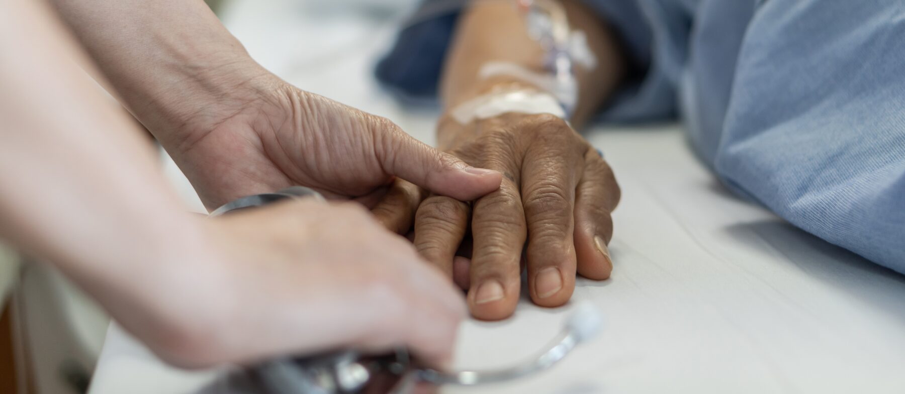 Gros plan sur la main d'un patient âgé alité. Une personne du corps médical lui tient la main.